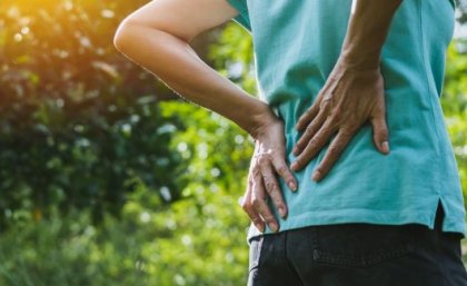 A person clutches their lower back as if in pain. They are outside with greenery in the background.